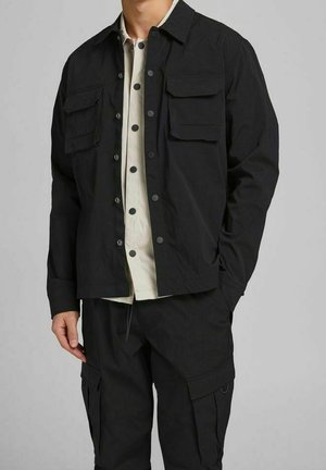 Person trägt eine schwarze, mit Knöpfen versehene Jacke mit Klappentaschen über einem teilweise zugeknöpften hellen Hemd und passenden schwarzen Cargohosen.