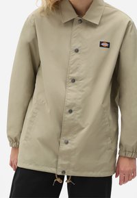 Světle béžová nepromokavá bunda s límečkem, patentkami, elastickými manžetami, bočními kapsami a značkovou nášivkou.