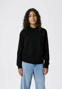 Felpa nera con cappuccio, dal fit rilassato, con polsini e orlo a costine. Abbinata a jeans a gamba larga di colore blu chiaro. Design minimalista su uno sfondo neutro.