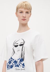 Hvit bomull t-skjorte med blått blomsterdesign og svart linjekunstillustrasjon av en kvinne med solbriller, rund halslinje og korte ermer.