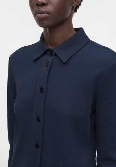 Personne portant une chemise bleu marine foncé à col avec boutonnière, montrée des épaules au menton sur un fond uni.