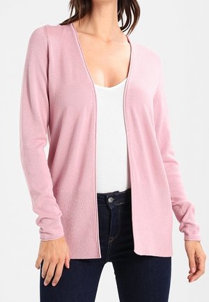 Frau trägt eine hellrosa offene Strickjacke über einem weißen Top und dunkelblauen Jeans, dargestellt vom Hals bis zu den Hüften.