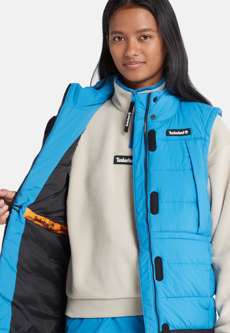 Blaues isoliertes Weste über einem cremefarbenen Fleece, mit schwarzen Akzenten und Taschen. Das Innere zeigt orangefarbenes Branding und eine glatte Textur.