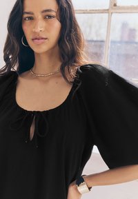 Sort, løstsiddende top med pufærmer og nøglehul med bindebånd i halsen. Glat stof med blød tekstur. Guldarmbånd på håndleddet.