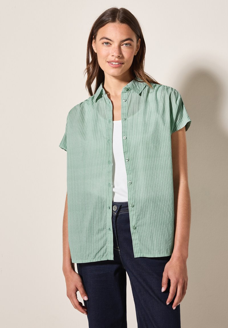 Camicia a maniche corte di colore verde chiaro con bottoni e righe verticali, caratterizzata da una texture morbida e un colletto classico. Indossata sopra una canotta bianca.