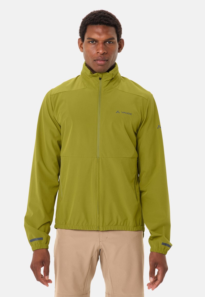 Homme portant une veste verte à fermeture éclair avec un col montant et un pantalon beige, debout devant un fond clair uni.