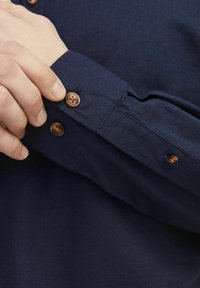 Camicia button-up blu navy con maniche in tessuto testurizzato, caratterizzata da due bottoni marroni sui polsini e una finitura liscia.