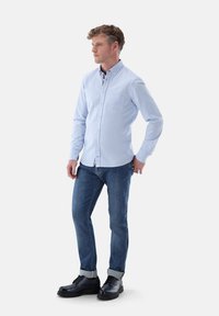 Hellblaues Button-up-Hemd mit einer Brusttasche, kombiniert mit dunkelblauen Jeans und schwarzen Schnürschuhen. Glattes Material und hochgerollte Ärmel.