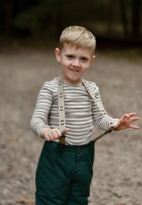 Niño rubio joven al aire libre, vestido con una camisa de manga larga a rayas, pantalones verdes con tirantes beige, sosteniendo un tirante con cada mano.