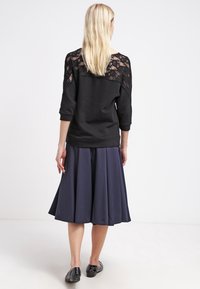 Felpa nera con pannello in pizzo sulle spalle, abbinata a una gonna svasata blu navy e scarpe flat nere. Texture morbide e vestibilità rilassata.