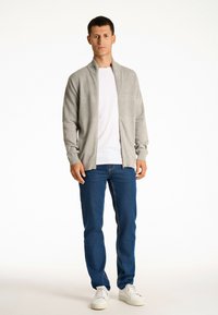 Helles graues Zip-Up-Cardigan mit strukturierten Einsätzen, getragen über einem weißen T-Shirt, kombiniert mit blauen Jeans und weißen Turnschuhen.