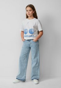 Weißes T-Shirt mit blauem Kirschgrafik und dem Text "CHERRYLICIOUS". Hellblaue weit geschnittene Jeans, weiße Sneaker. Einfaches, lässiges Outfit.