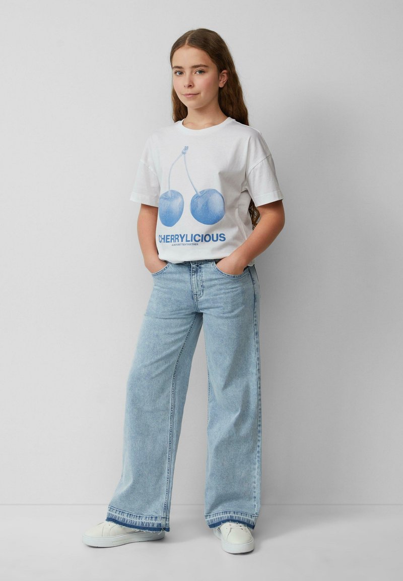 Weißes T-Shirt mit blauem Kirschgrafik und dem Text "CHERRYLICIOUS". Hellblaue weit geschnittene Jeans, weiße Sneaker. Einfaches, lässiges Outfit.