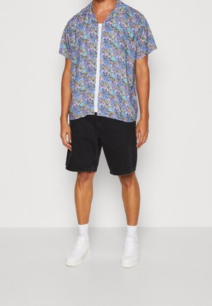 Camicetta a maniche corte blu con un motivo astratto colorato, abbinata a pantaloni corti in denim neri e sneakers bianche.