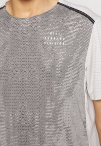 Grå og hvid Nike løbetrøje med vandrette striber, rund halsudskæring, korte ærmer og teksten "Nike Running Division" i hvid.