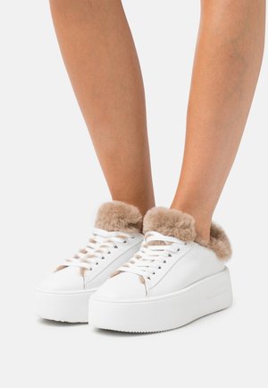 Baskets à plateforme blanches avec doublure en fourrure beige portées sur des jambes nues sur un fond blanc uni.