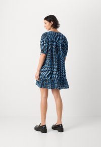 Abito a maniche corte con motivo geometrico blu e nero, caratterizzato da un orlo a balze e una vestibilità ampia. La modella indossa delle scarpe con zeppa nere.