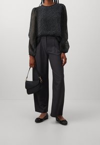 Svart mönstrad blus med genomskinliga, puffiga ärmar, kombinerad med högmidjade grå byxor. En liten svart handväska och svarta ballerinas fulländar outfiten.