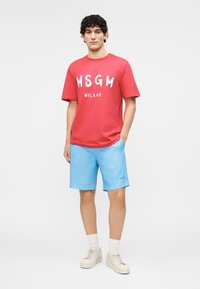 Jeune homme debout portant un T-shirt rouge MSGM Milano, un short bleu clair, des chaussettes blanches et des baskets beiges sur un fond uni.