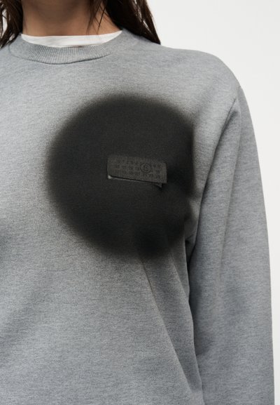 Άτομο που φοράει γκρι φούτερ με μαύρο κυκλικό λεκέ ή αποχρωματισμό στην αριστερή πλευρά του στήθους, καλύπτοντας εν μέρει μια μικρή υφασμάτινη ετικέτα.