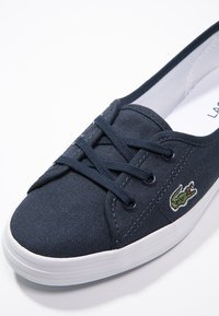 Baskets en toile bleu marine avec semelle en caoutchouc blanc, équipées de lacets ronds et du logo crocodile brodé sur le côté. Texture douce, design à bout arrondi.