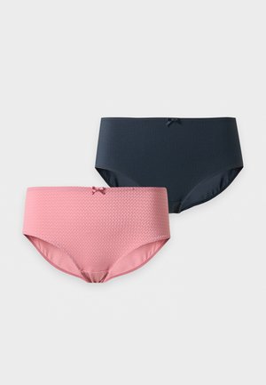 Mujer con cabello largo y oscuro que lleva un top de bikini marrón triangular y braguitas de bikini negras de talle alto, de pie contra un fondo claro y liso.