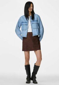 Moteris dėvi šviesiai mėlyną džinsinę striukę, baltą palaidinę, rudą zomšinę sijoną ir juodus iki kelių aukščio batus, stovinti su rankomis kišenėse prie balto fono.