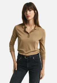 Polo à manches longues de couleur marron clair avec un col et des détails en bouton, associé à un jean noir taille haute et une ceinture noire.