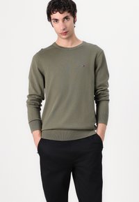 Pulover verde măsliniu dintr-un tricot moale, având guler rotund și manșete cu nervuri, purtat împreună cu pantaloni negri. Detaliu cu logo pe piept.