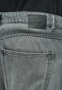 Jean en denim gris avec une étiquette de marque en cuir sur la ceinture, présentant une coupe classique, une poche arrière et des détails de couture subtils.