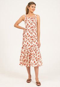 Robe midi florale avec décolleté carré, bretelles spaghetti, base crème, motif de fleurs rouges, détail de boutons et ourlet à volants ; associée à des sandales marron.