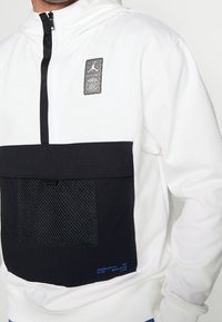 Sudadera blanca con capucha y un bolsillo de malla negra, con una cremallera negra y un parche de logo. El material parece suave con un acento texturizado.
