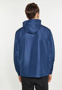 Marineblaue, wasserfeste Jacke mit Kapuze, die eine strukturierte Oberfläche und elastische Bündchen aufweist. Die Rückansicht zeigt ein schlichtes Design ohne auffällige Details.