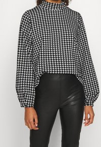 Haut à manches longues en motif pied-de-poule noir et blanc, avec un col montant, une coupe ample et des poignets froncés, associé à un pantalon en simili cuir noir.