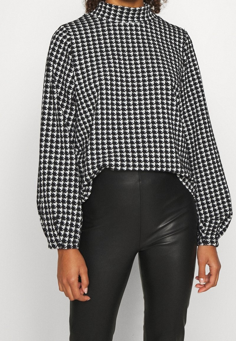 Haut à manches longues en motif pied-de-poule noir et blanc, avec un col montant, une coupe ample et des poignets froncés, associé à un pantalon en simili cuir noir.