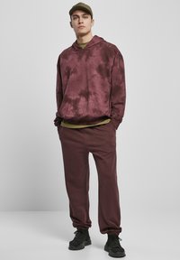 Hanorac tie-dye maroniu cu buzunar frontal, suprapus peste o cămașă verde măslin, asortat cu pantaloni jogger maronii și adidași negri.