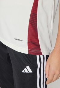 Balta sportinė marškinė su raudonais raštais šoniniu paneliu ir "AEROREADY" logotipu; suderinta su juodais šortais su balta Adidas trijų juostų dizainu.