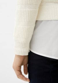 Gebreide trui met witte strepen, met een geribde manchet. Onder de trui is een witte hemdzoom zichtbaar, gecombineerd met donkere jeans.