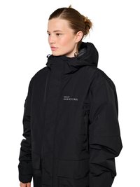 Vaaleaihoinen nainen, jolla on ruskeat hiukset nutturalla, pukeutuneena mustaan huppariin, jossa on teksti "HALO 1968|972|981" rinnassa, katsoo vasemmalle.