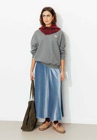 Sweatshirt gris superposé sur un sweat à capuche bordeaux, associé à une longue jupe bleu satinée, des chaussures à lacets marron et un sac fourre-tout vert olive.