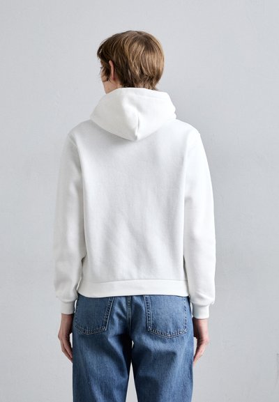 Sweat-shirt à capuche blanche avec une texture lisse, des poignets ajustés et un ourlet côtelé, associé à un jean en denim bleu. Design et coutures visibles au dos.