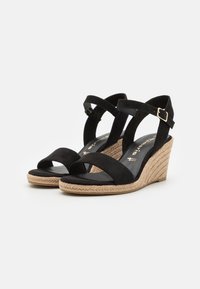 Sandales compensées noires avec brides à la cheville et boucles dorées, dotées de talons tissés style espadrille et orteils ouverts.