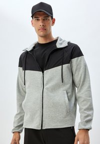 Grau und schwarze Kapuzenjacke mit Reißverschluss, mit einem Kordelzug an der Kapuze, seitlichen Taschen und einer weichen Textur. Das schwarze Vorderteil kontrastiert mit dem grauen Körper.