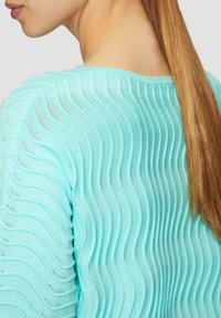 Pull en tricot bleu clair avec un motif en vagues, présentant des lignes texturées et un col rond. Les cheveux sont lisses et tombent dans le dos.