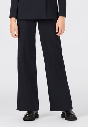 Osoba nosząca czarne, szerokie spodnie w cienkie prążki, czarną kurtkę i czarne masywne skórzane buty stojąca na jasnoszarej podłodze.