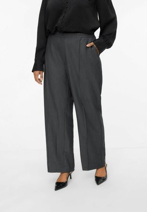 Frau trägt dunkelgraue, weit geschnittene Hose, schwarze Bluse mit Knöpfen und schwarze spitze High Heels, steht mit einer Hand in der Tasche.