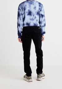 Niebiesko-biała sweter tie-dye, czarne sztruksowe spodnie z kieszeniami oraz szare tenisówki z żółtymi akcentami. Ujęcie od tyłu.