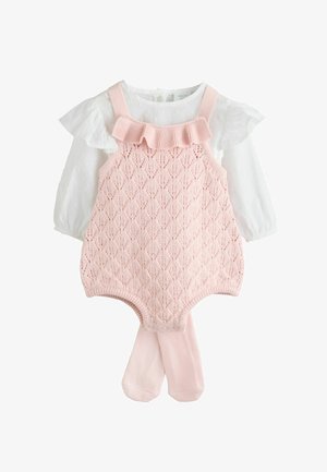 Ružový pletený overal s textúrovaným diamantovým vzorom, ktorý je doplnený o bielu blúzku s volánikmi a zodpovedajúce ružové pančuchy.