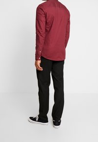 Muž stojící zády, oblečený v dlouhém tmavě červeném tričku, černých kalhotách a černých teniskách Converse s bílými tkaničkami.