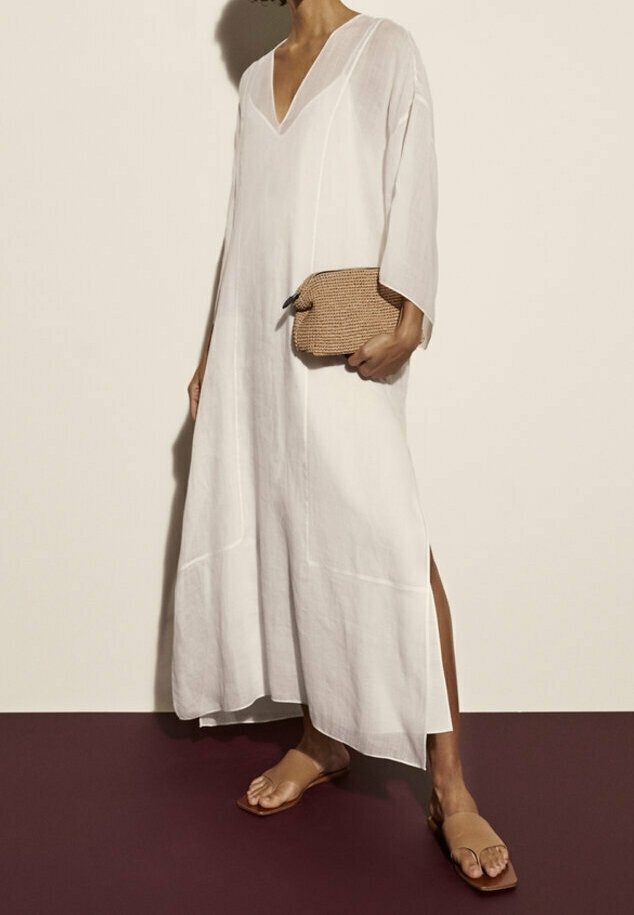 Femme portant une robe blanche ample à manches longues avec des fentes latérales, tenant une pochette tissée et chaussant des sandales slides marron clair sur un sol bicolore.